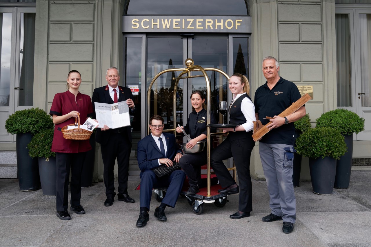 Mitarabeitende aus verschiedenen Abteilungen vom Hotel Schweizerhof Luzern posieren lachend vor dem Haupteingang des Hotels.