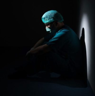 Medizinischer Mitarbeitender sitzt erschöpft am Boden einer dunklen Ecke und wird beleuchtet