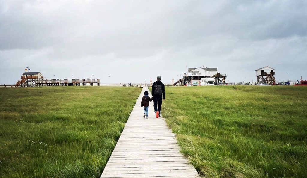 Ich gehe mit meinem Sohn an der Hand einen langen Holzsteg entlang. Im Hintergrund ist der Strand der Nordsee sichtbar.