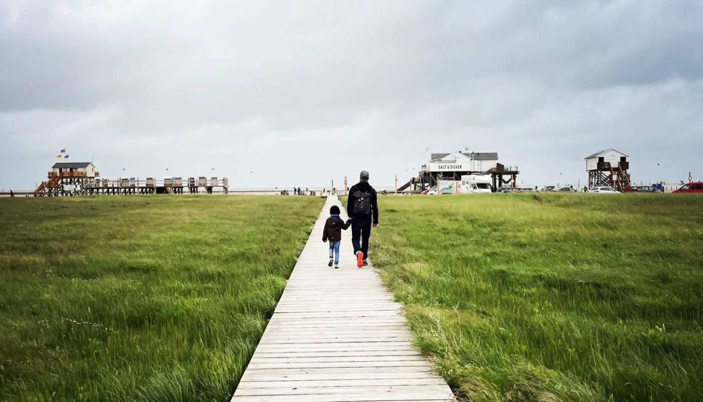 Ich gehe mit meinem Sohn an der Hand einen langen Holzsteg entlang. Im Hintergrund ist der Strand der Nordsee sichtbar.