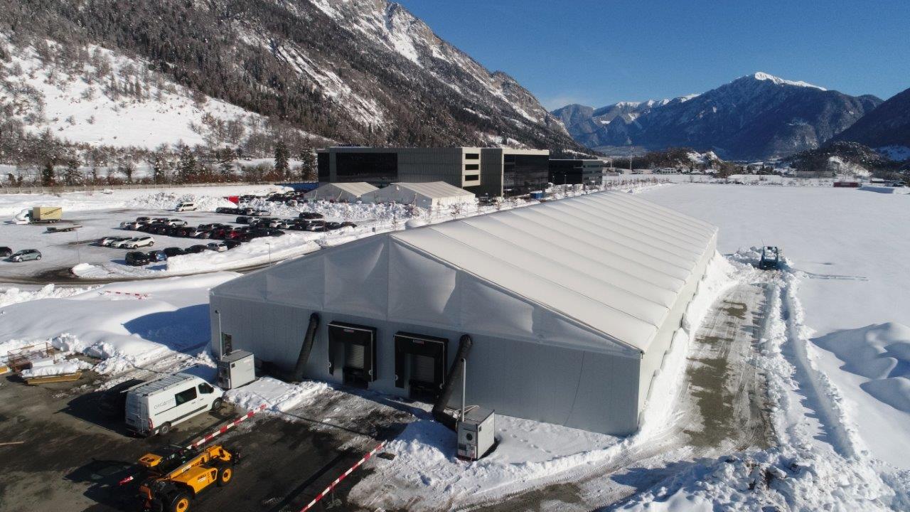 Luftaufnahme einer mobilen Industriehalle von Orgatent im Wintereinsatz: Die weiße Halle mit transluzenter Thermo-Dachplane und isolierten Seitenwänden steht auf einem geräumten, asphaltierten Gelände vor schneebedeckten Alpen im Hintergrund. Zwei Verladerampen mit Andocktoren sind an der Stirnseite installiert. Umgeben von Schnee, Fahrzeugen und einer angrenzenden Gewerbezone zeigt das Bild die Flexibilität und Einsatzfähigkeit temporärer Hallenlösungen unter extremen Wetterbedingungen. 