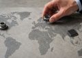 Eine Hand hält ein Zahnrad über einer Weltkarte. Ein Halbleiterchip liegt auf Asien und ein Metallbauteil auf Nordamerika.