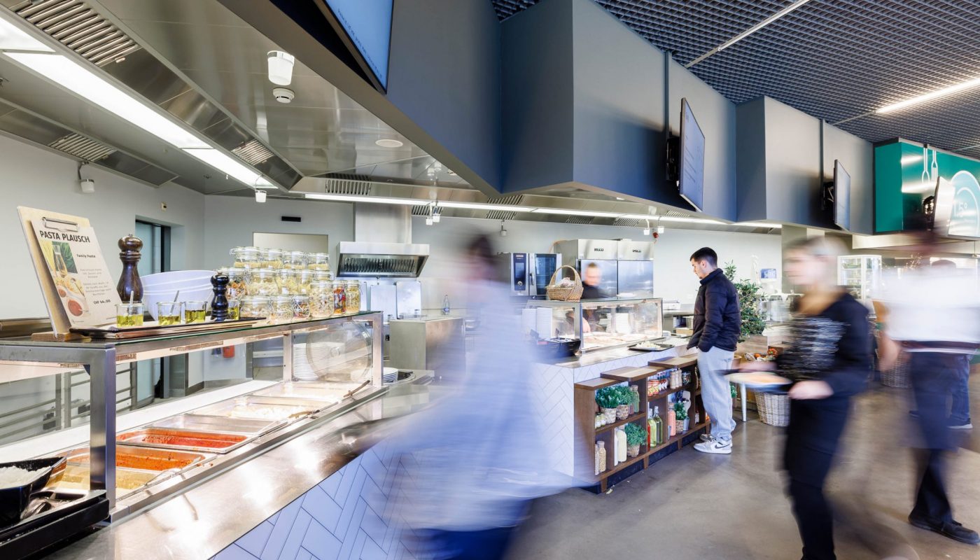Mehrere Gäste bewegen sich durch ein modernes Selbstbedienungsrestaurant mit offenen Ausgabestationen und Buffettheken.