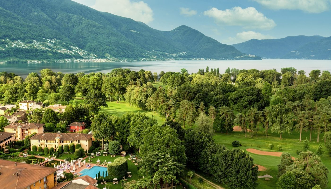 Red and orange buildings, surrounded by nature such as trees and lawn. Alongside of a golf court. Hills and the Lago Maggiore are in the back visible.