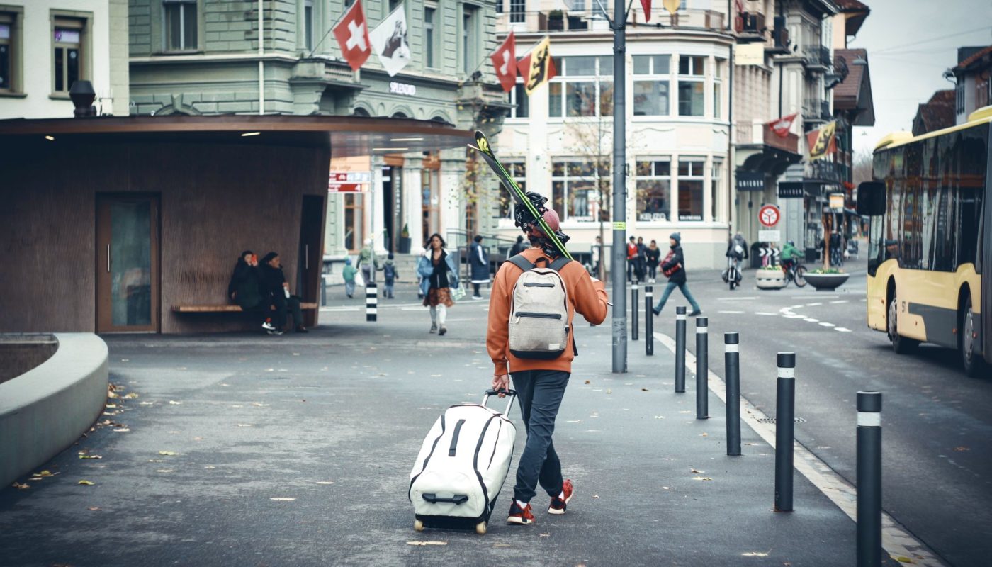 Mann mit Koffer und Ski auf der Strasse einer Stadt in der Schweiz. Im Hintergrund Menschen und Flaggen (Schweiz, Bern).