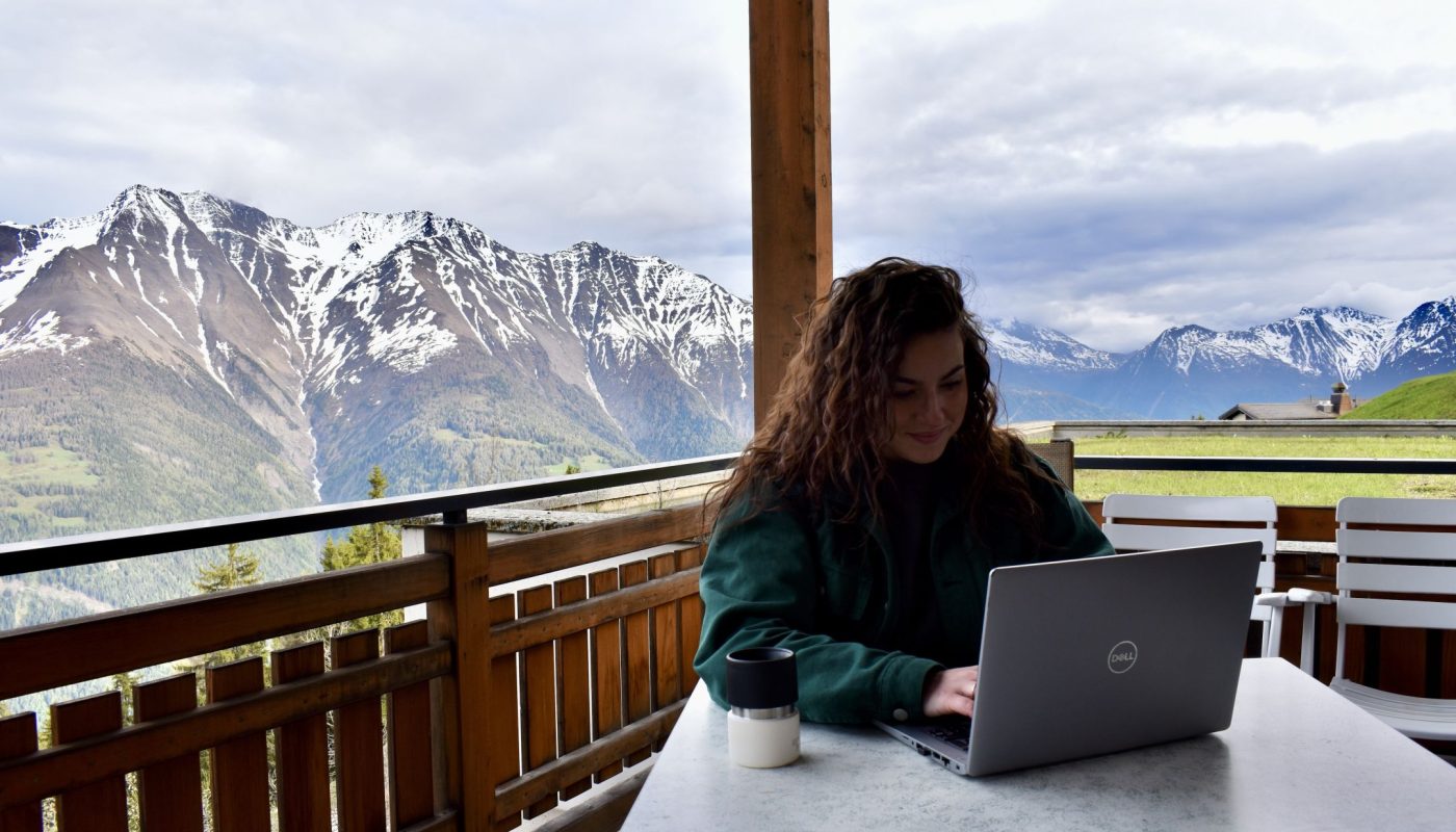 Junge Frau arbeitet auf einem Balkon am Laptop auf der Riederalp. Im Hintergrund Walliser Bergkette.
