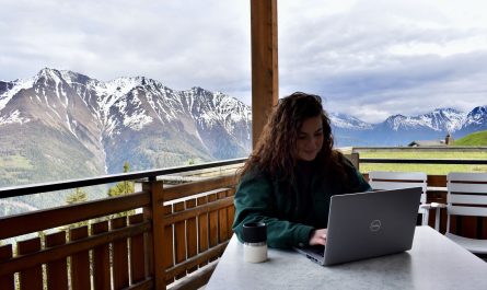 Junge Frau arbeitet auf einem Balkon am Laptop auf der Riederalp. Im Hintergrund Walliser Bergkette.