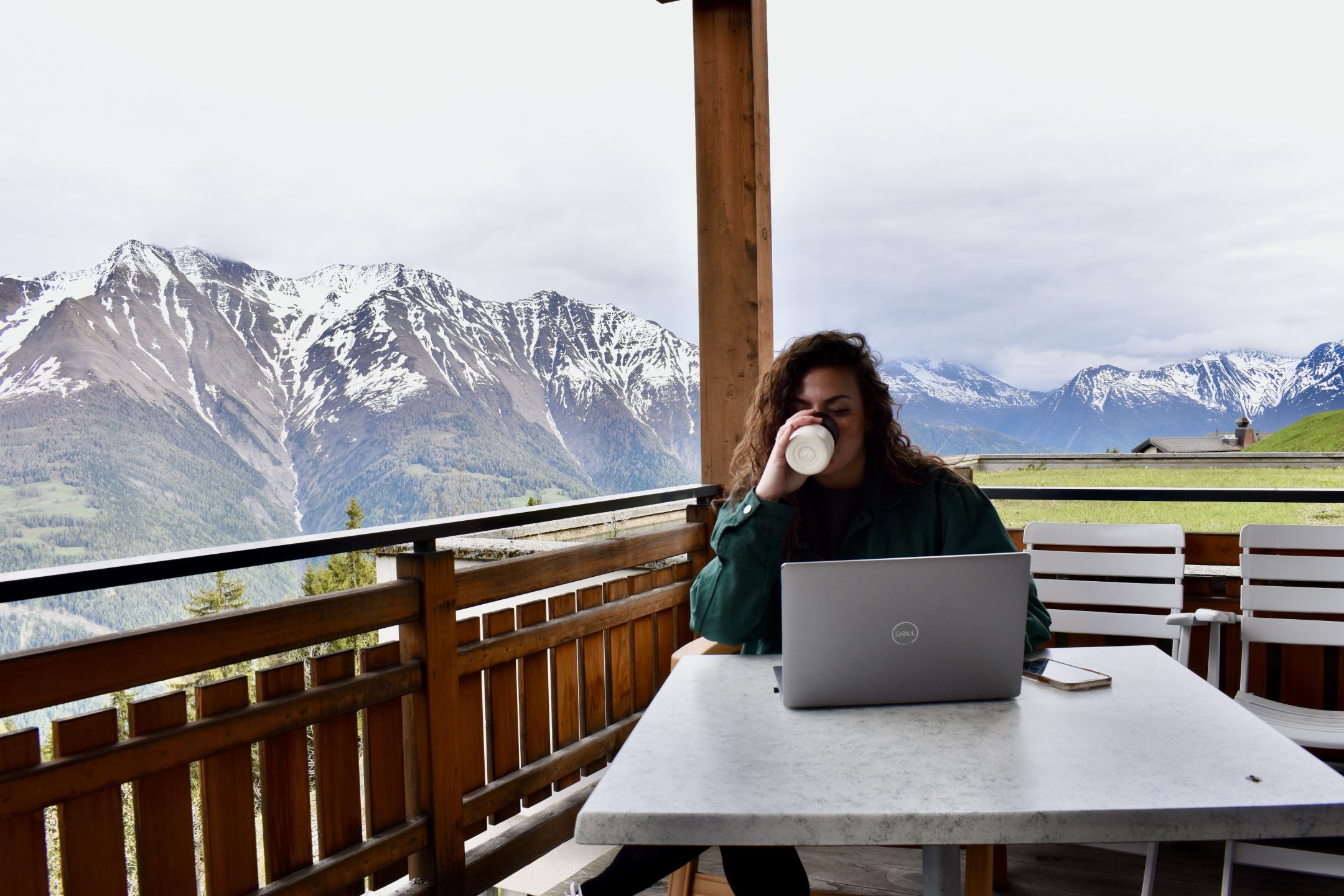Junge Frau arbeitet auf einem Balkon am Laptop und trinkt Kaffee. Im Hintergrund Walliser Bergkette. 