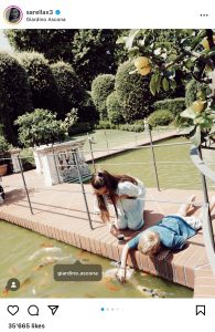 A woman and a child feeding Koi fish. Kneeling on the bridge of the water lily pond. Surrounded by water, trees and lemon trees.