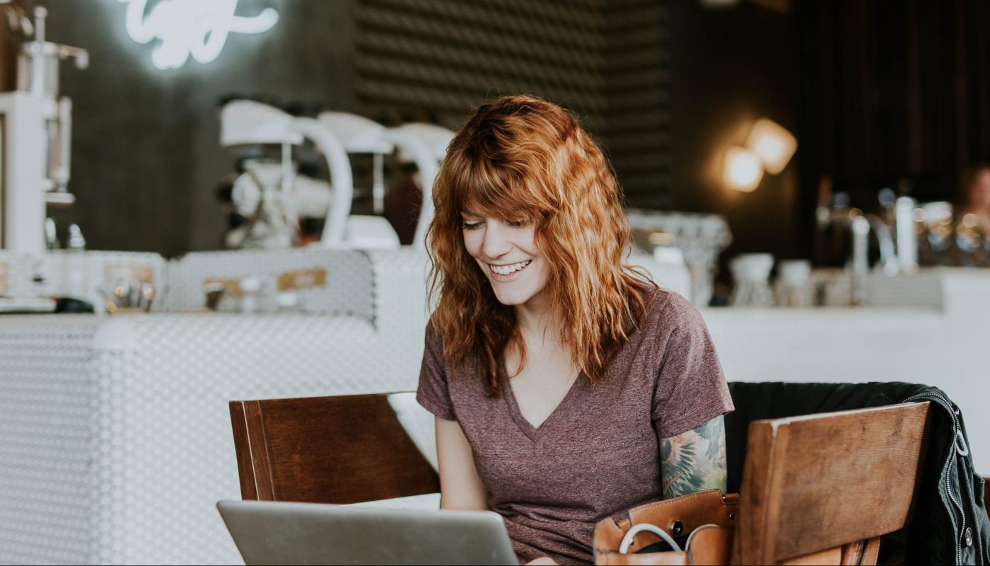 Junge, rothaarige Frau lächelt und arbeitet am Laptop in einem gemütlichen Café