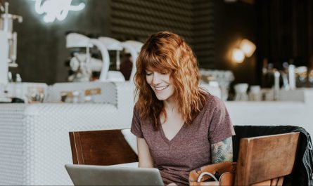 Junge, rothaarige Frau lächelt und arbeitet am Laptop in einem gemütlichen Café