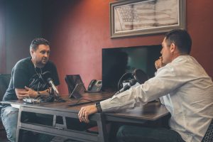 Zwei Männer, welche gemeinsam am Tisch sitzen und einen Podcast aufnehmen