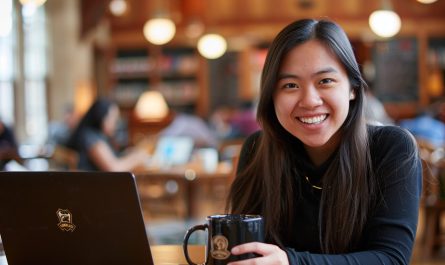 A happy university student sits at a library table with a laptop, holding a coffee mug. The background suggests a focused yet relaxed academic environment with other students studying.