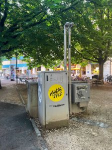 Community fridge from Madame Frigo in Helvetiagärtli, Lucerne, promoting food sustainability and waste reduction. The public refrigerator is placed in an outdoor setting with trees and nearby cafes, allowing locals to share surplus food and support a sustainable community effort.
