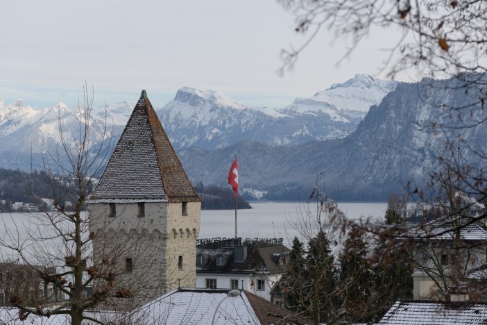 View of a historic tower in Lucerne with a Swiss flag flying, set against a wintry backdrop of Lake Lucerne and snow-covered Alps. Bare tree branches frame the scene, highlighting the peaceful winter atmosphere of central Switzerland.