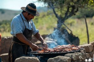 Gaucho grilling traditional Argentinian asado outdoors over open fire in rural countryside.