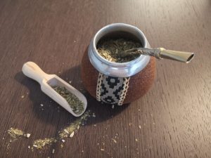 Traditional Argentine mate gourd filled with yerba mate and a metal bombilla straw, placed on a wooden table with a wooden scoop and loose yerba leaves beside it—representing the cultural ritual of drinking mate in Argentina.